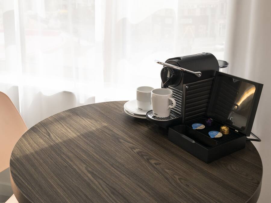 Una mesa redonda de madera con una máquina de café, dos tazas blancas y una bandeja con una caja pequeña y un objeto azul.