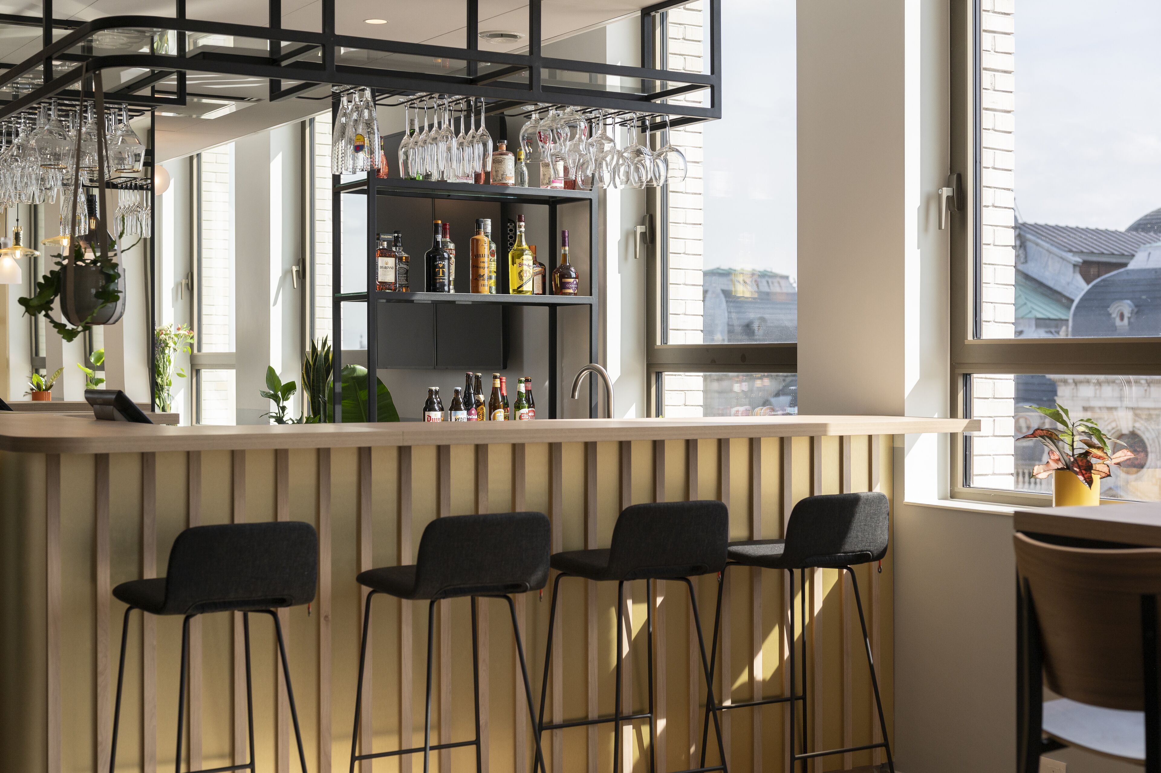 Un coin bar avec trois tabourets noirs, une étagère remplie de bouteilles et de verres, et une fenêtre donnant sur des immeubles.