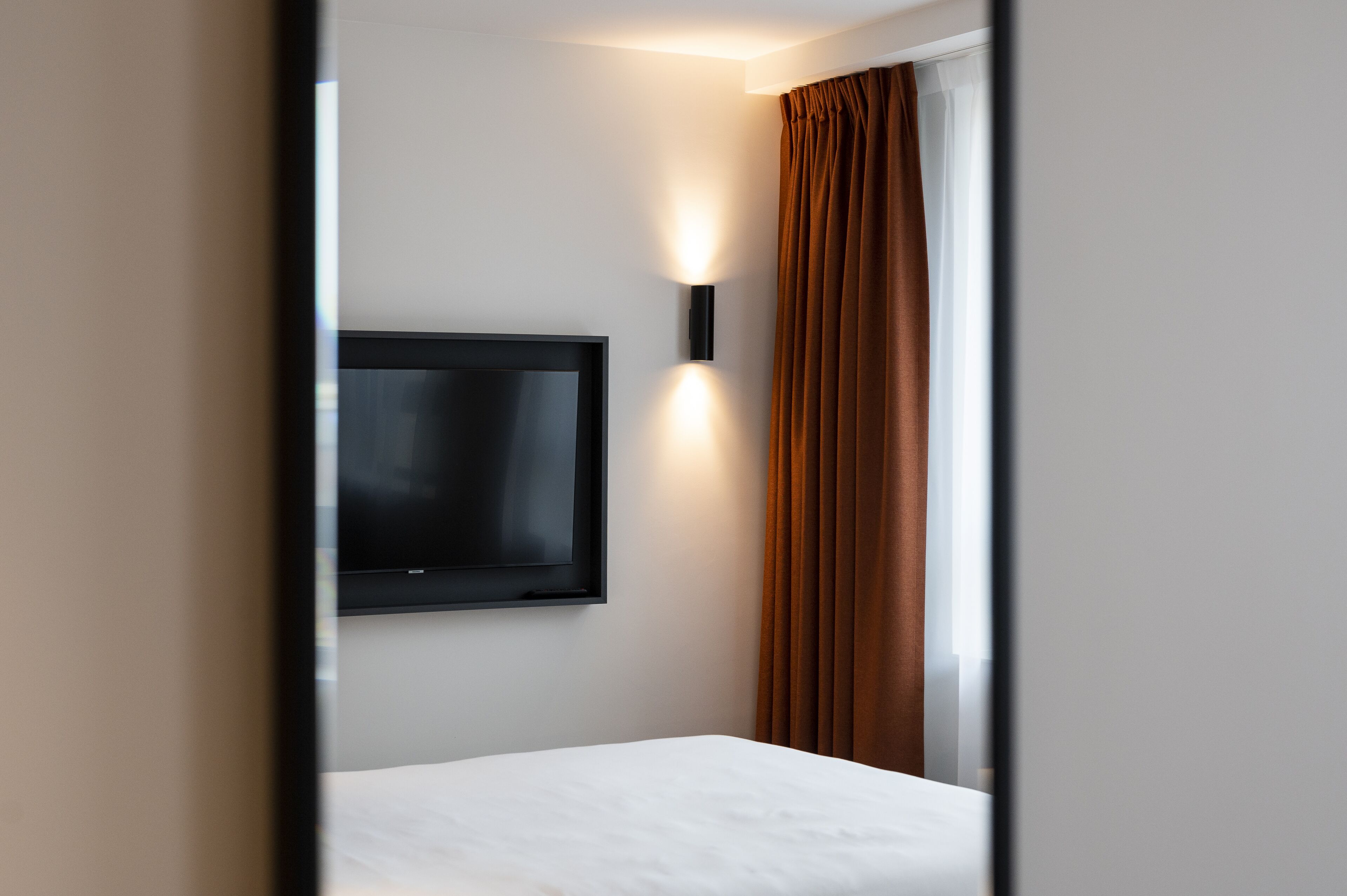 A hotel room with a flat-screen TV mounted on the wall, a window with brown curtains, and a white bed.