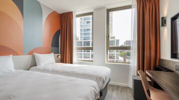 Habitación de hotel con dos camas, un escritorio y una silla. Amplias ventanas con cortinas ofrecen vistas de edificios cercanos.
