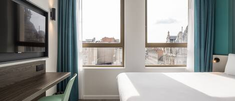 A hotel room with a bed, a desk, a chair, a TV, and large windows with curtains.