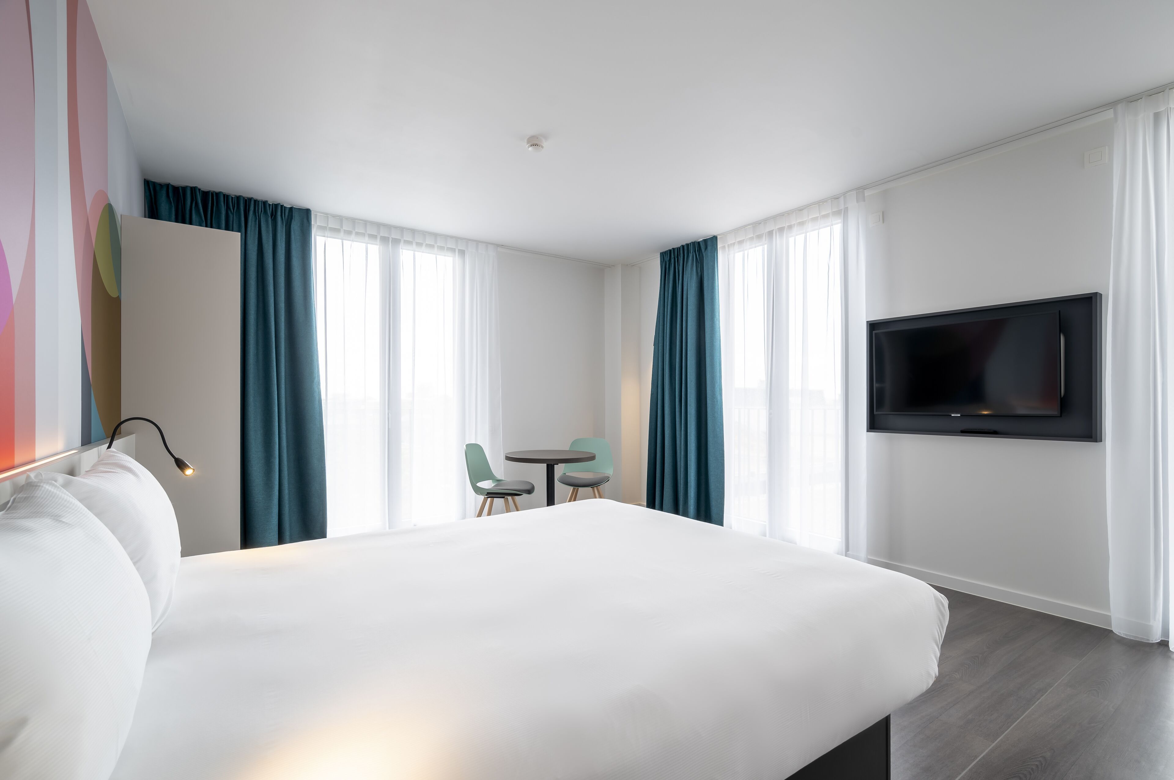 Une chambre d’hôtel avec un grand lit, un téléviseur fixé au mur, une petite table avec deux chaises et un bureau avec une chaise.