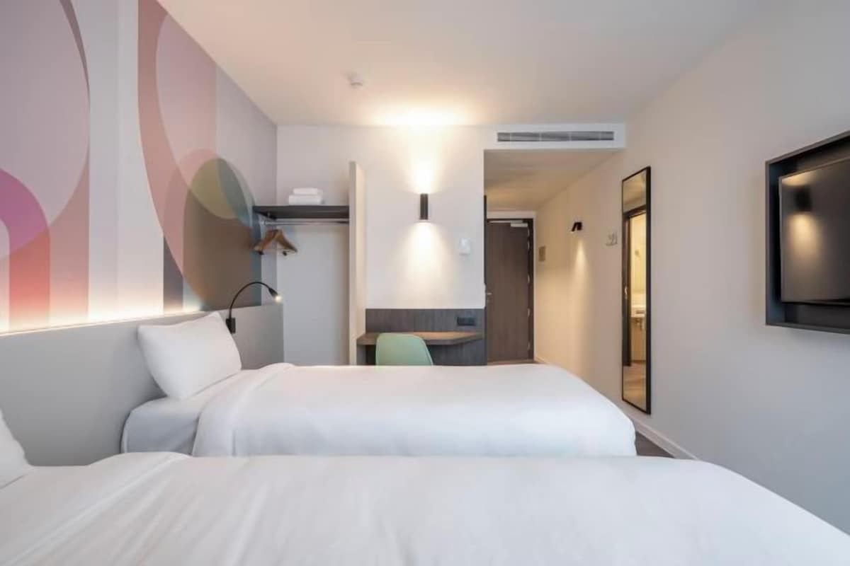 A hotel room with two beds, a desk, a chair, a TV, and a wall with abstract art.
