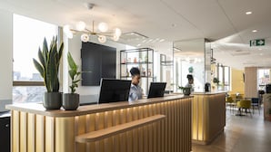 A modern reception desk with a wooden finish, two potted plants, and a person behind the counter. - B&B HOTEL Antwerpen Centrum (Antwerp)