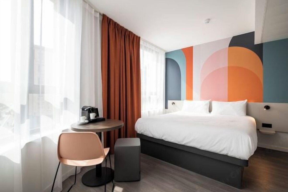 A modern hotel room with a bed, a round table with a coffee maker, a chair, and a small ottoman.