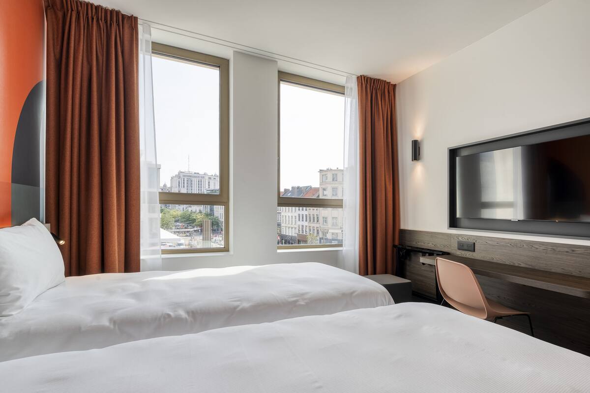Habitación de hotel con dos camas, televisión y ventanas amplias con cortinas.