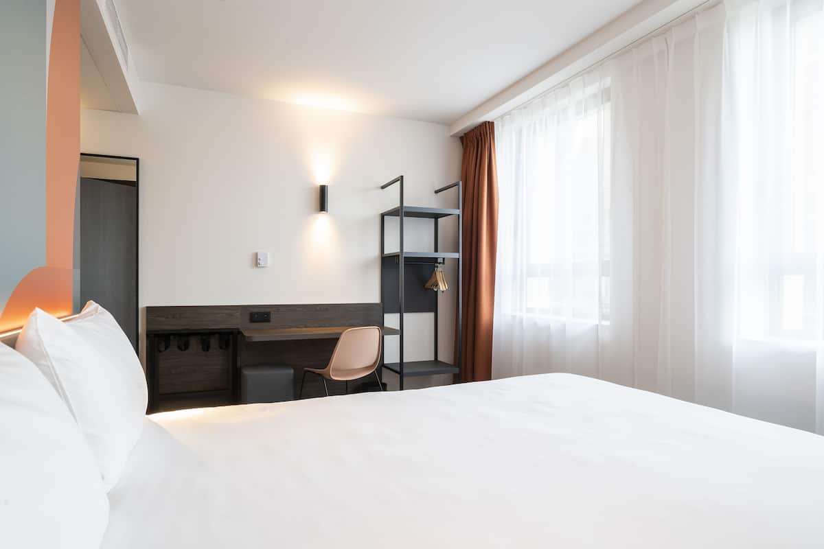 Habitación de hotel con cama, escritorio, silla, estante y lámpara de pared.