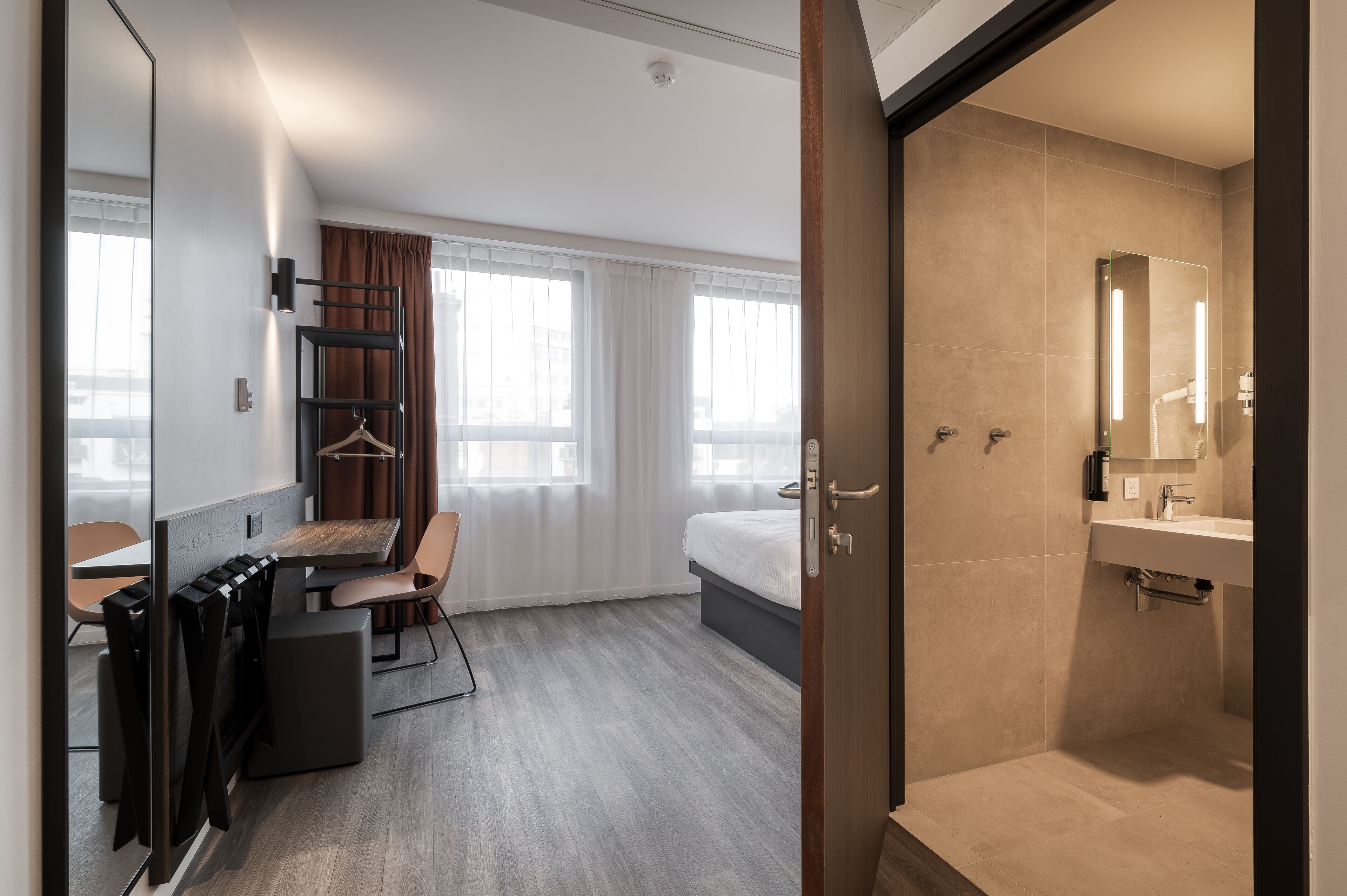 Een moderne hotelkamer met een bed, een bureau met stoel, een kledingkast en een badkamer met wastafel en spiegel.