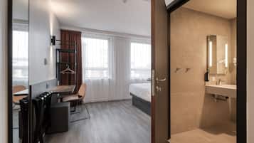 Een moderne hotelkamer met een bed, een bureau met stoel, een kledingkast en een badkamer met wastafel en spiegel.