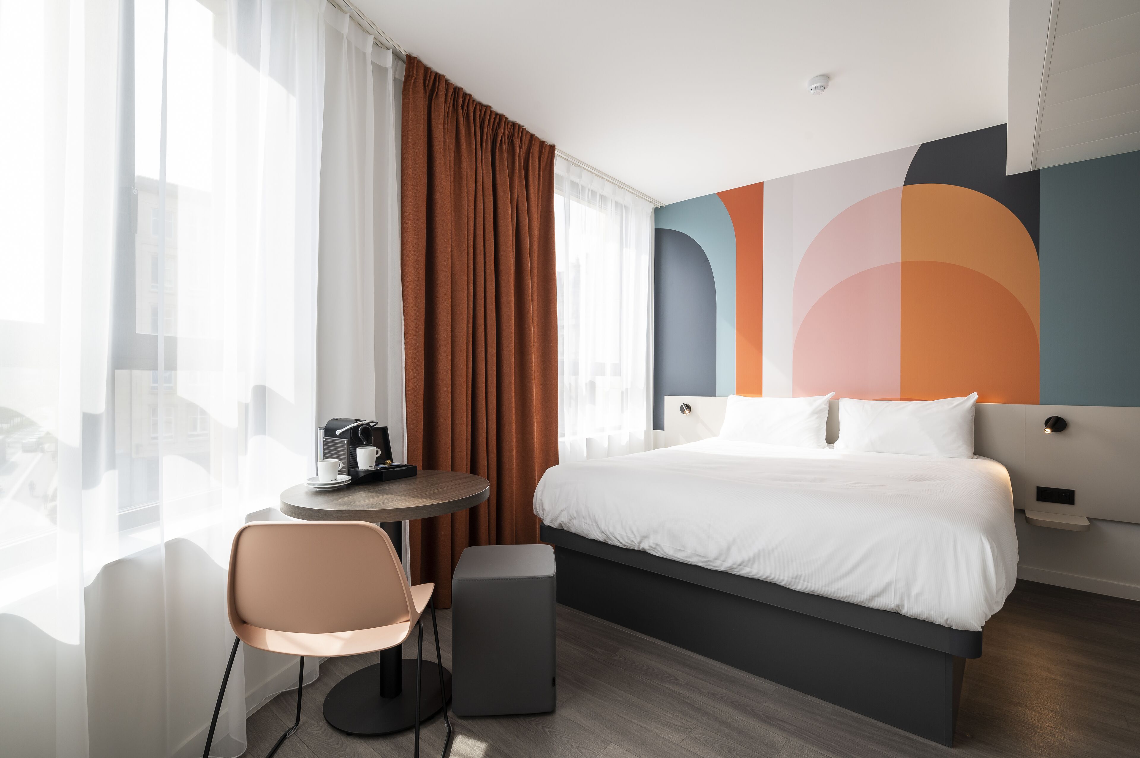 Una habitación de hotel moderna con una cama grande, una mesa redonda con cafetera, una silla y vistas de la ciudad a través de cortinas translúcidas.
