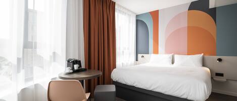 Una habitación de hotel moderna con una cama grande, una mesa redonda con cafetera, una silla y vistas de la ciudad a través de cortinas translúcidas.