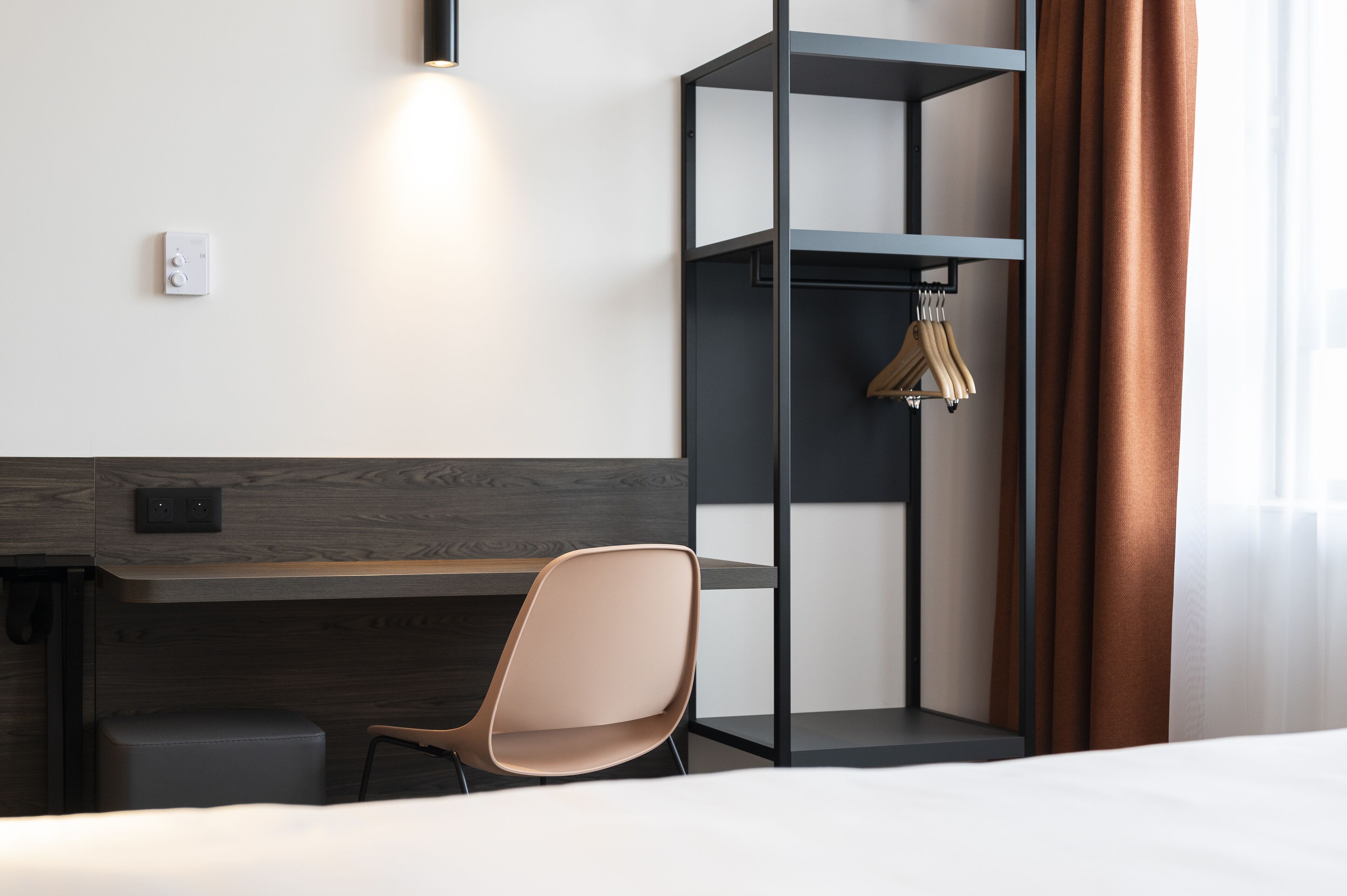 A modern hotel room with a wooden desk, a cushioned chair, a wardrobe, and a hanging coat rack.