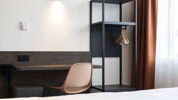 A modern hotel room with a wooden desk, a cushioned chair, a wardrobe, and a hanging coat rack.