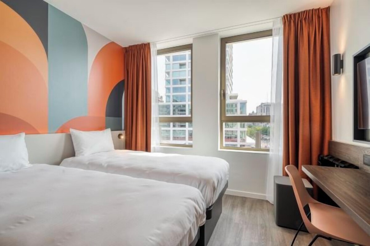 A hotel room with two beds, a desk, and a large window with curtains.