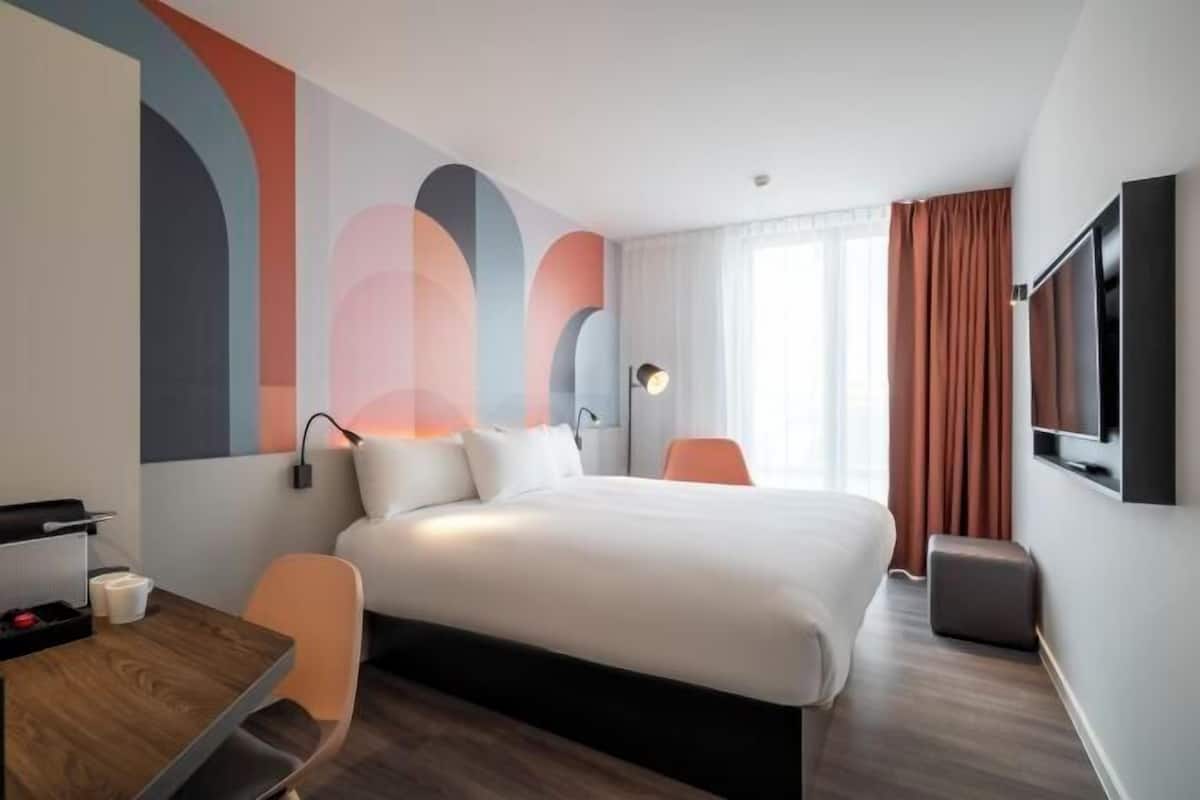 Una habitación de hotel moderna con una cama grande, un escritorio, una silla, un televisor y una pared con arte abstracto.
