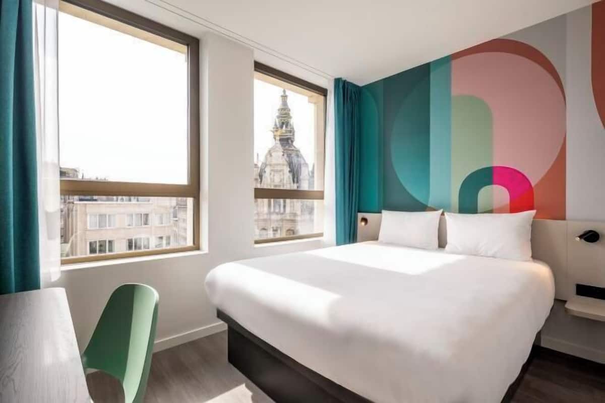 Habitación de hotel con una cama grande, dos ventanas, vista a un edificio y un diseño de pared colorido.