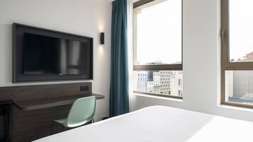 Hotelkamer met een bed, een aan de muur bevestigde televisie, een bureau met stoel, en een raam met uitzicht op gebouwen.