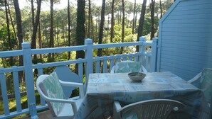 A covered outdoor seating area with a table and chairs, surrounded by a white railing, overlooking a forested area. - In the middle of the pines, attractive apartment for 2 - 4 people (Lacanau)