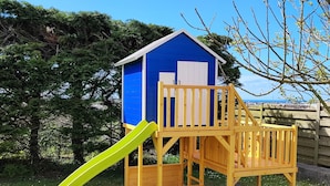 Een houten speelhuisje met een blauwe buitenkant en een gele glijbaan.
