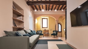 Ein gemütliches Wohnzimmer mit einer Couch, Sesseln und einem Fernseher. Regale mit Büchern und ein Fenster mit Tageslicht.