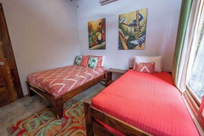 A room with two wooden beds, each with a red bedspread. There are two paintings on the wall, one depicting birds and the other a tropical scene. A window with curtains is visible on the right.