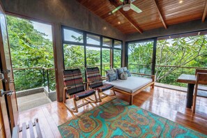 A spacious room with a wooden ceiling, large windows, a wooden floor, a patterned rug, and a seating area with chairs and a bench.