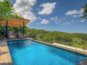 A swimming pool with a wooden pergola and lounge chairs, surrounded by lush greenery and a clear sky.