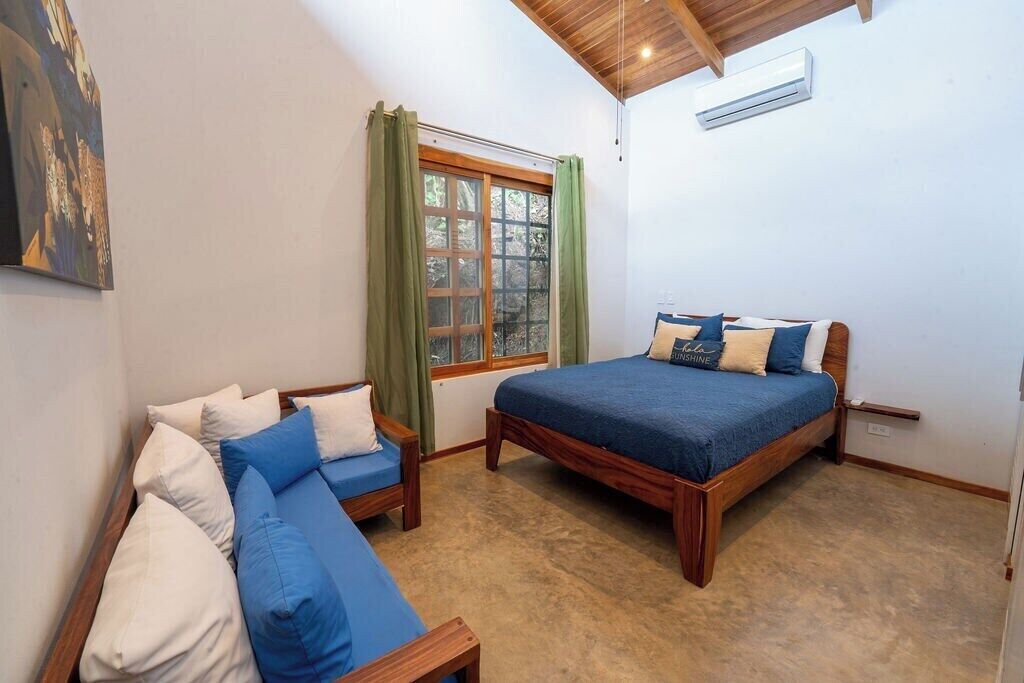 A bedroom with a bed, a sofa, a window with curtains, and an air conditioning unit.