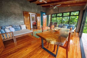 A spacious room with a wooden dining table, a sofa, and a large window offering a view of greenery.