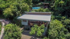 A two-story building surrounded by dense greenery, with a visible balcony on the upper floor.