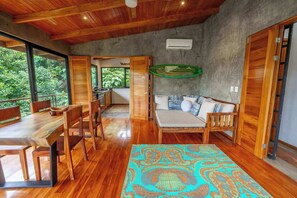 A spacious room with a wooden dining table and chairs, a sofa with cushions, and a large window offering a view of greenery.