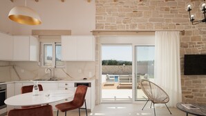 A modern kitchen with white cabinets, a dining area with a white table and brown chairs, and a large sliding glass door leading to an outdoor patio with a pool and seating. - Evaero Villa II, Family Retreat with a 32m² Pool (Prinos)
