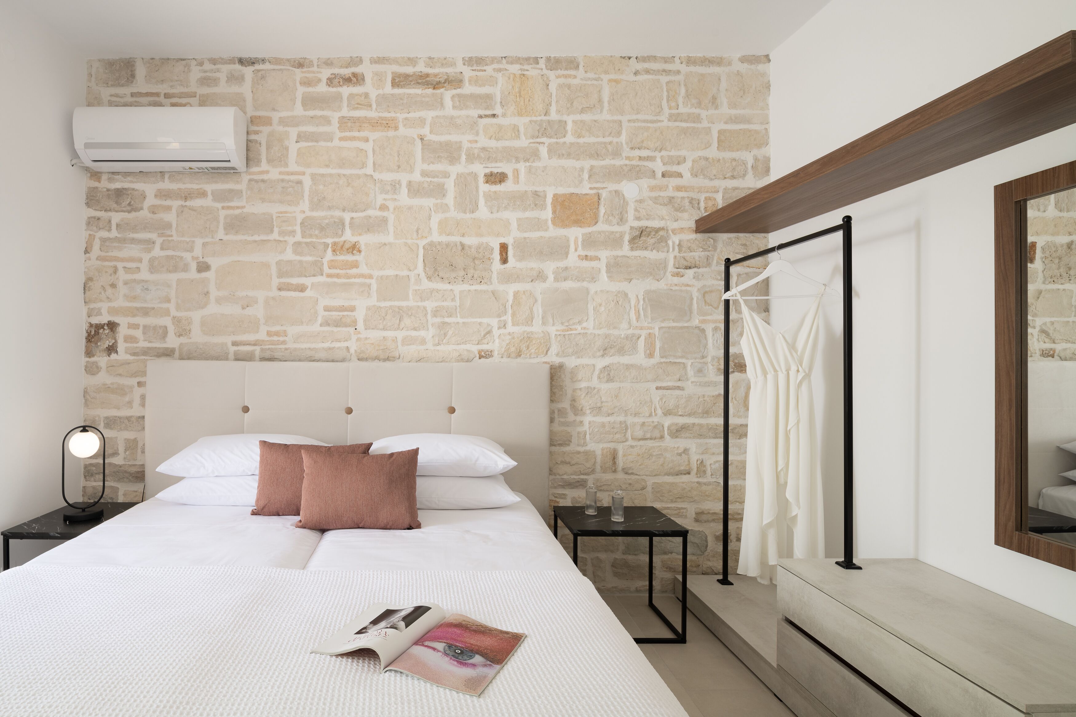 Una camera da letto con parete in pietra, un letto con biancheria bianca, un comodino con una lampada e una finestra con tende.