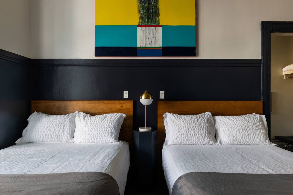 A hotel room with two beds, a wooden headboard, a modern painting, and a nightstand with a lamp.