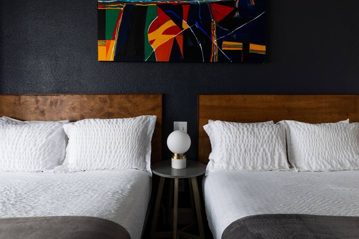 A hotel room with two beds, a round table with a lamp, and a colorful abstract painting on the wall.