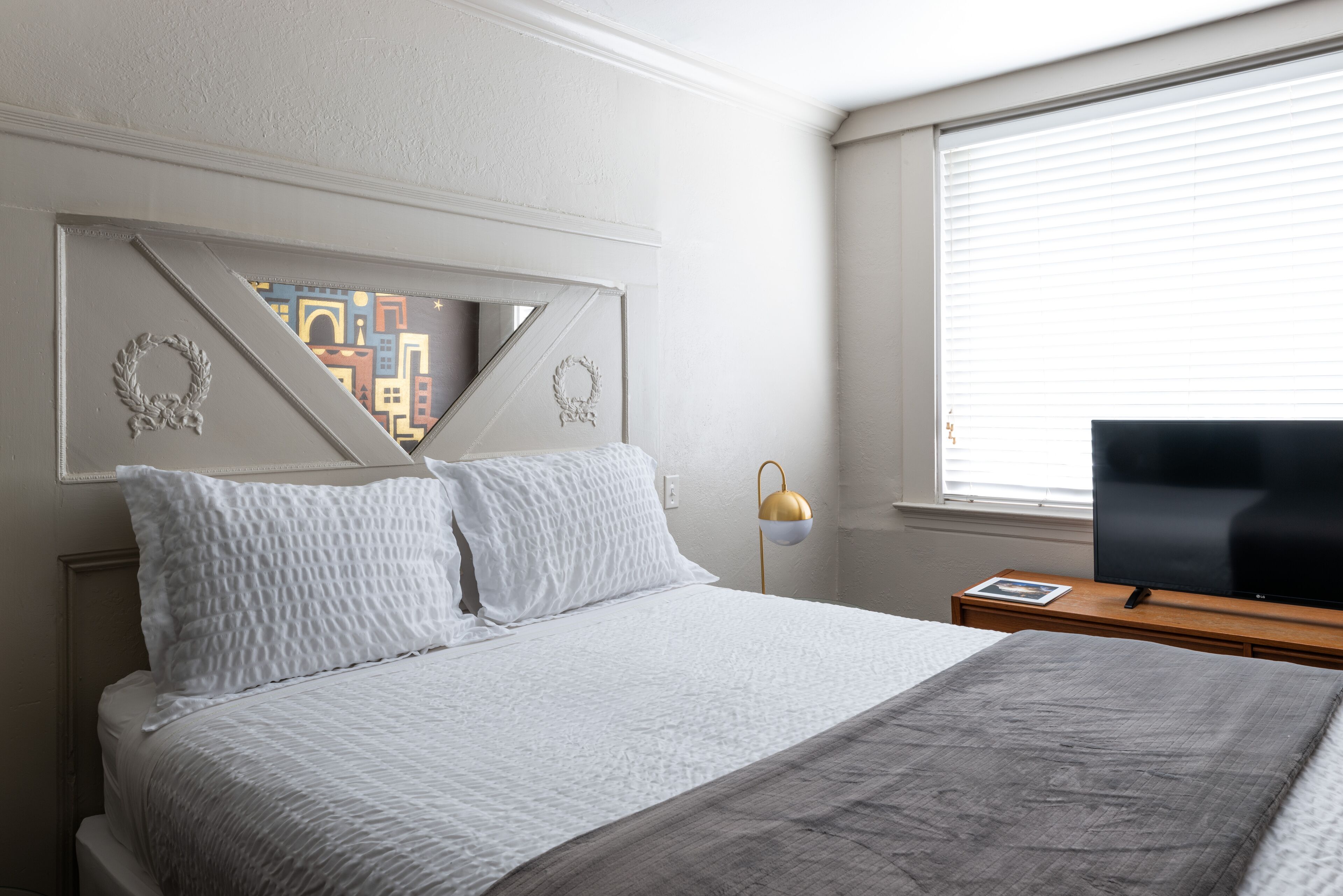 A neatly made bed with white linens, a bedside table with a lamp, and a TV on a wooden stand.