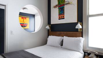 A neatly made bed with white linens, a round mirror on the wall, and a painting above it.