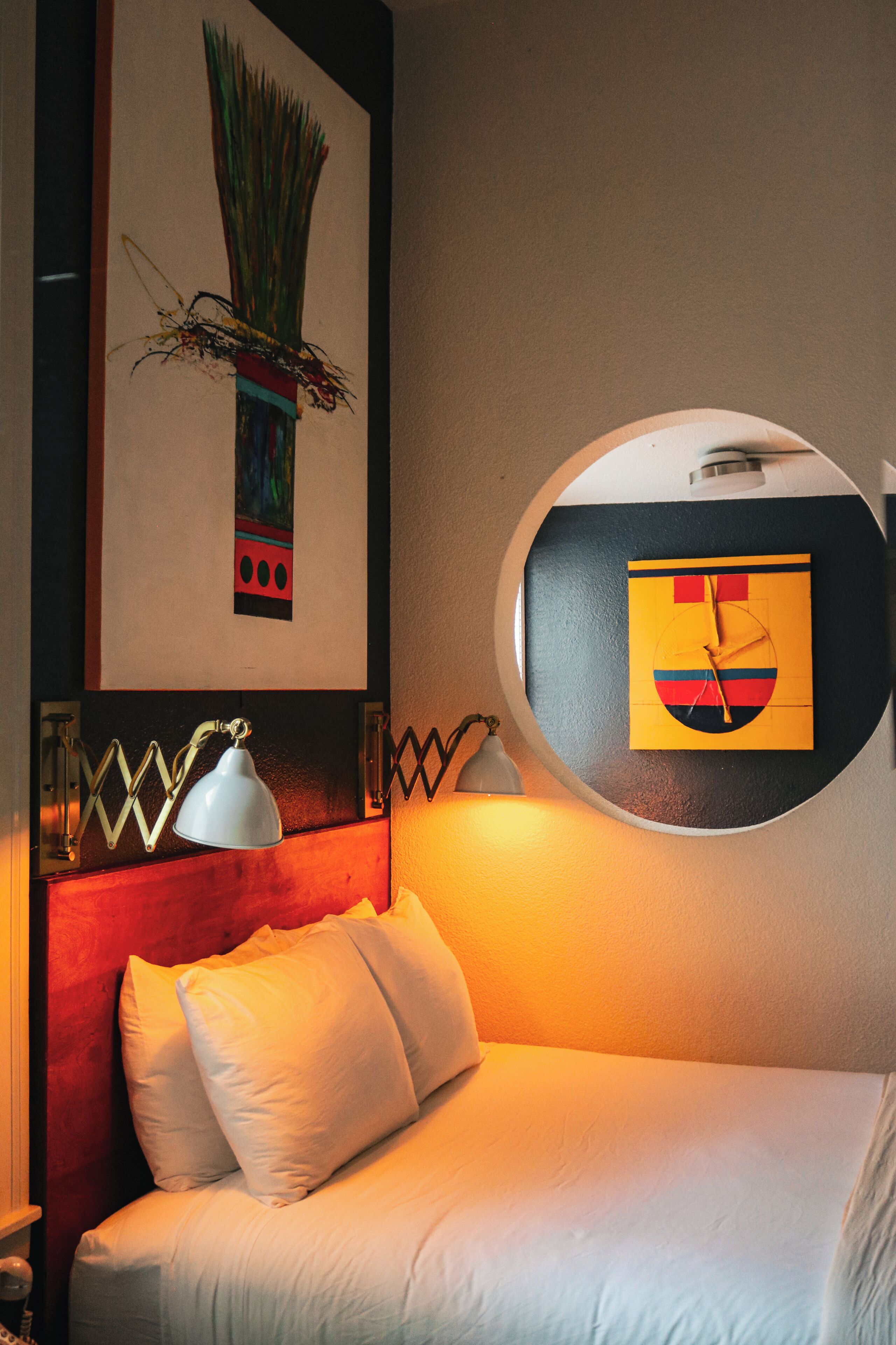 A hotel room with a bed, a round mirror, a wall clock, and a framed artwork above the bed.