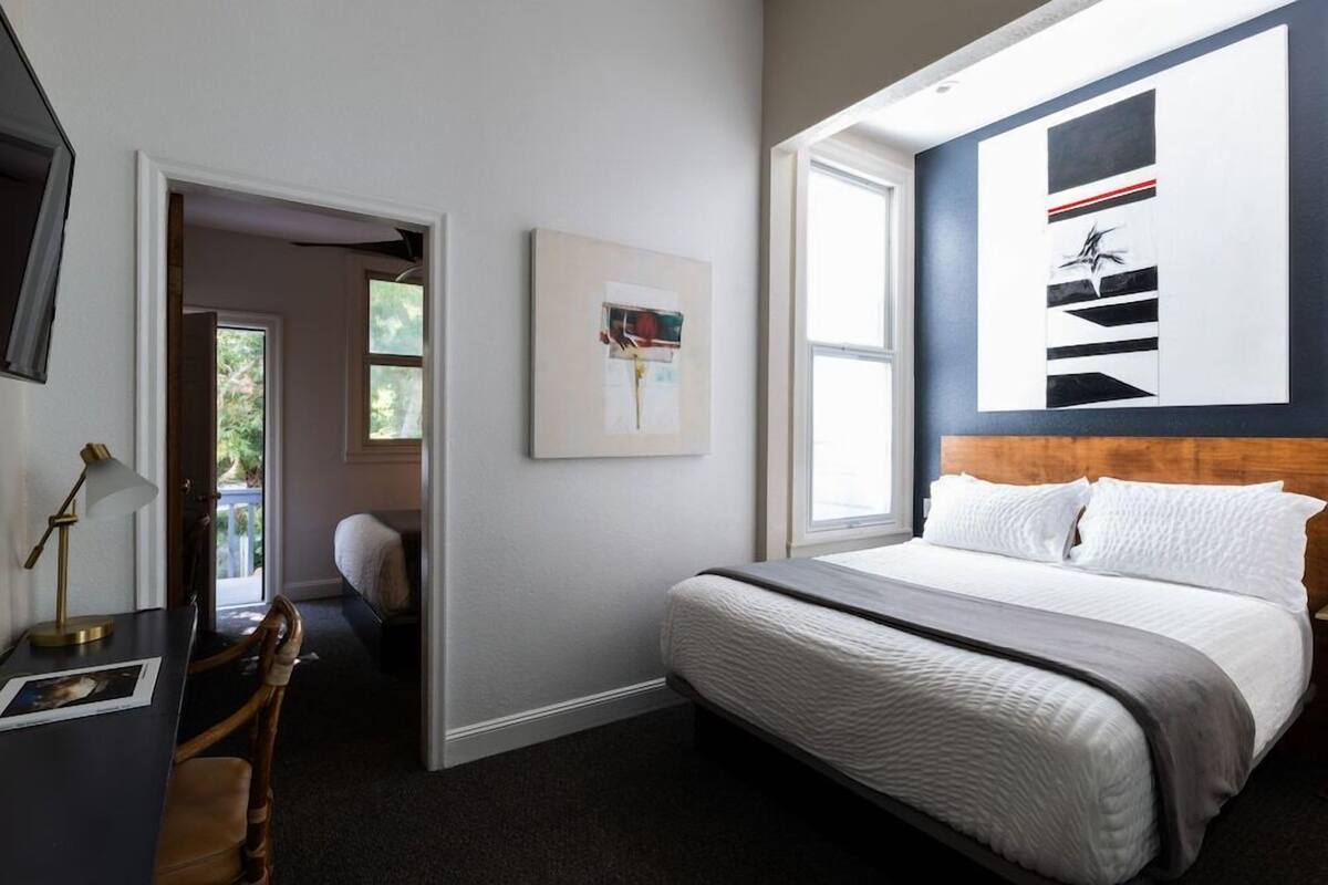 A bedroom with a bed, a desk with a lamp, a chair, a window, and artwork on the wall.