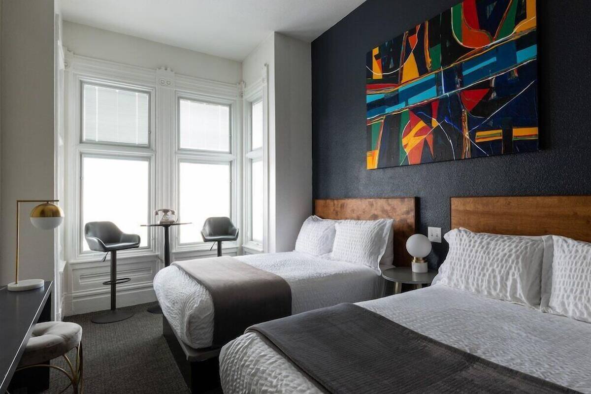 A hotel room with two beds, a large abstract painting, a window with curtains, a small table, and a chair.