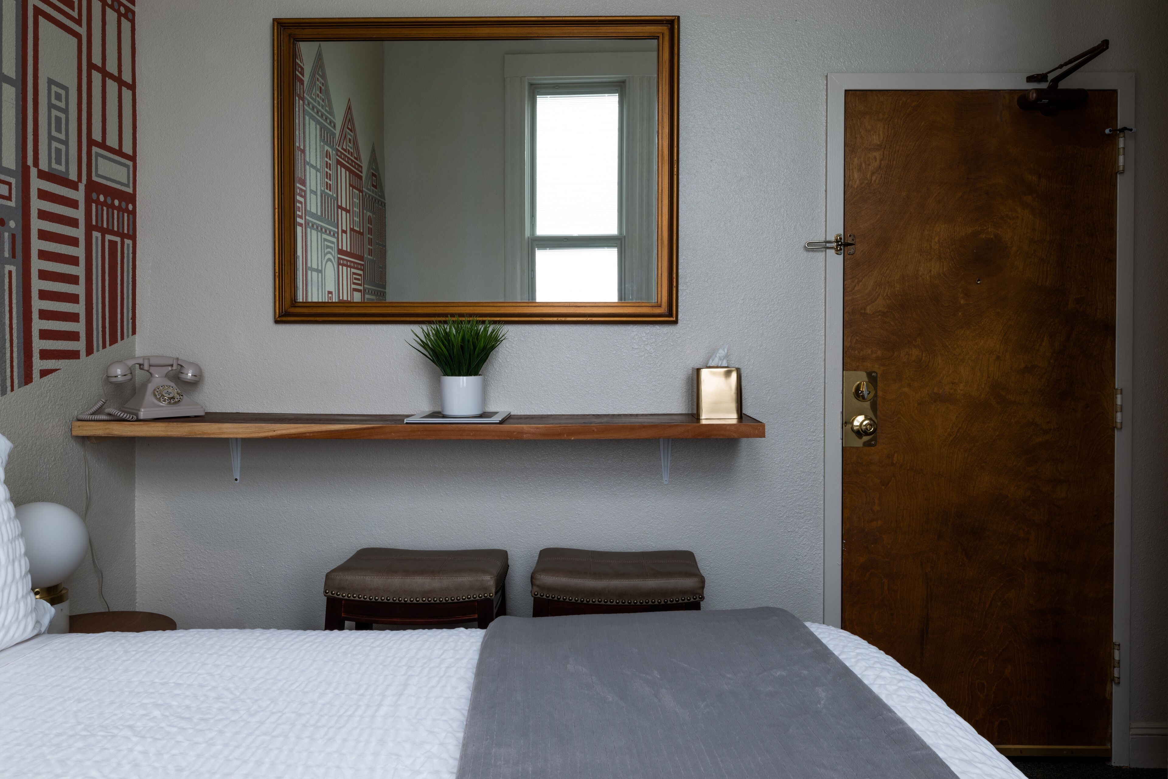 A bedroom with a large bed, a wooden door, a mirror, a shelf with a plant, and a telephone.