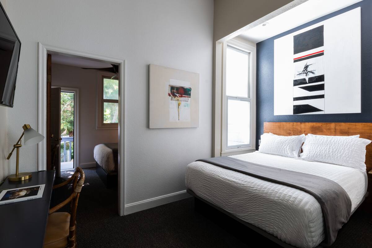 A bedroom with a bed, a desk with a lamp, a chair, a painting on the wall, and a window.