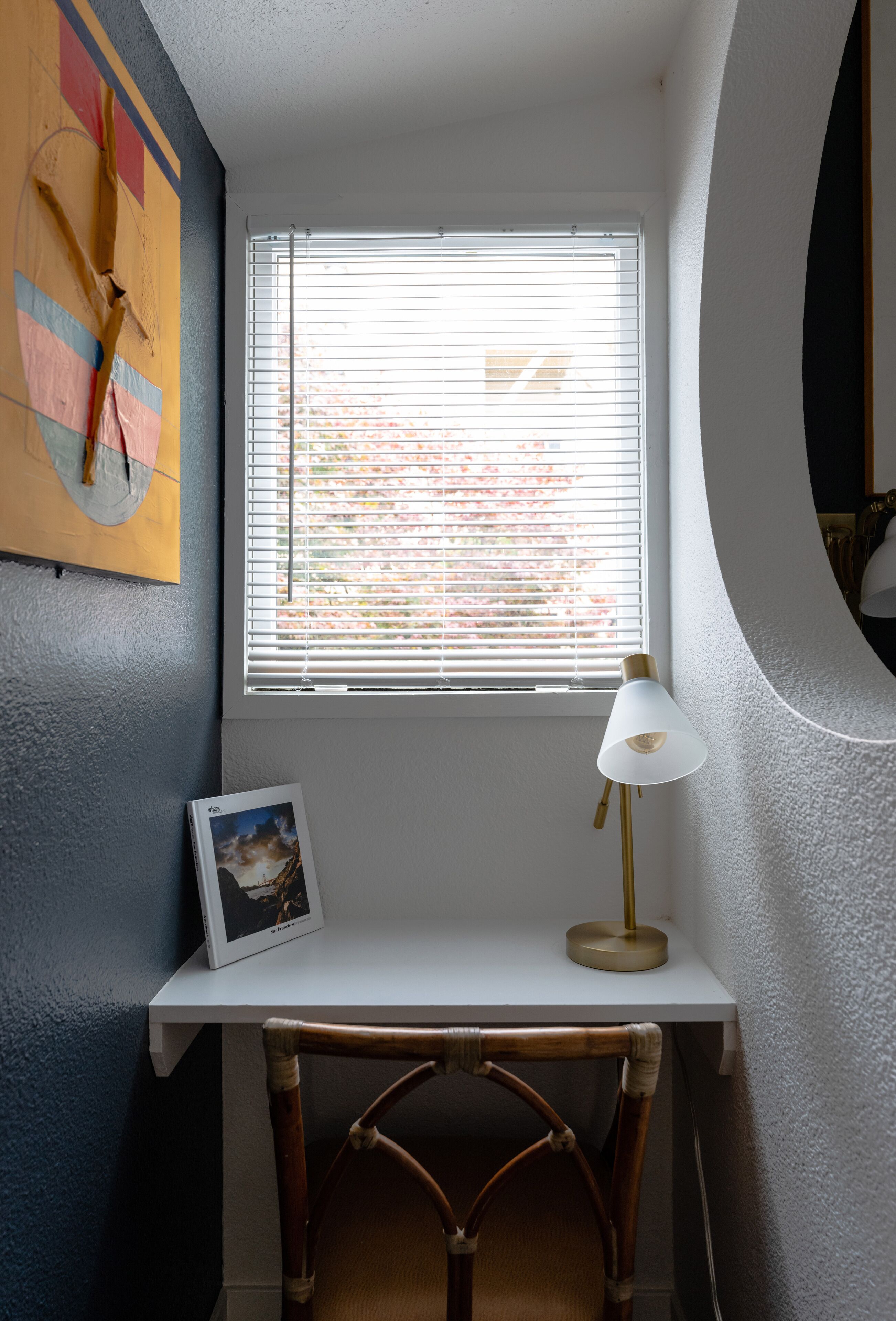 A room with a desk, a chair, a window with blinds, a lamp, and a framed picture on the wall.
