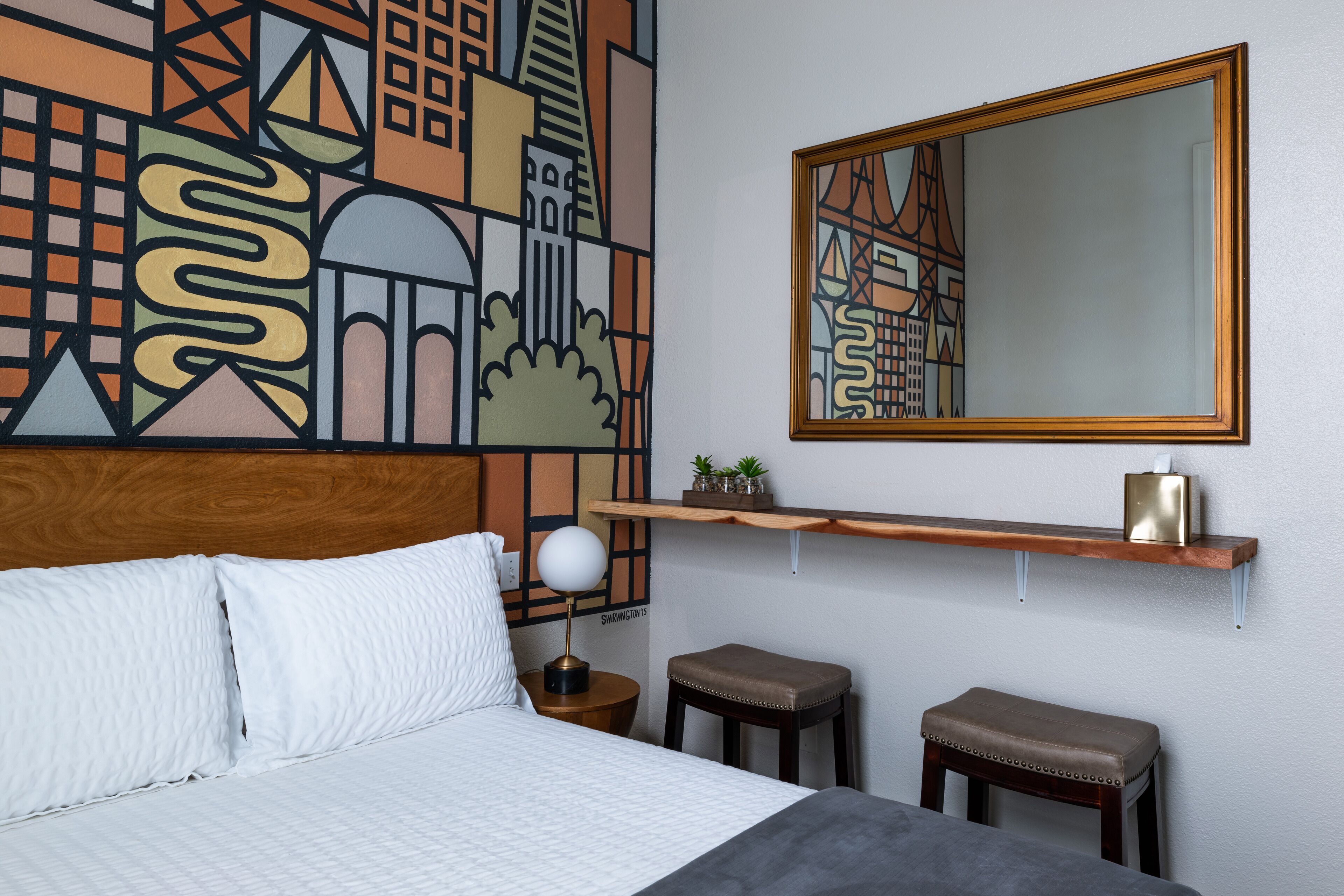 A hotel room with a bed, a nightstand with a lamp, two stools, a wooden shelf with a tissue box, and a large abstract cityscape mural on the wall.