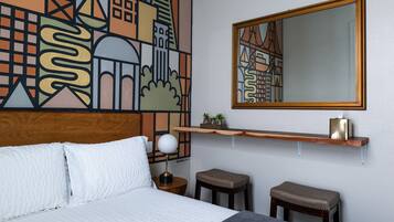 A hotel room with a bed, a nightstand with a lamp, two stools, a wooden shelf with a tissue box, and a large abstract cityscape mural on the wall.