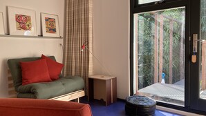 A modern living room with a green sofa, red cushions, a small wooden side table, and a black ottoman. There are two framed artworks on the wall, a floor lamp, and a sliding glass door leading to a balcony with a view of greenery. - Houseboat with private studio near downtown and Rai in the middle of urban greenery (Amsterdam)