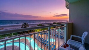 Un balcon avec une piscine et des chaises longues, offrant une vue sur une plage et l’océan au coucher du soleil.
