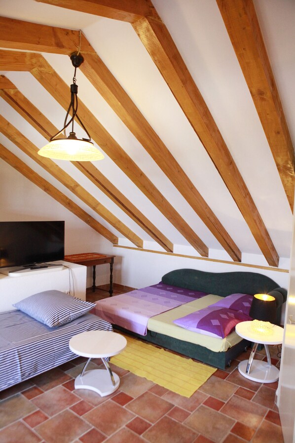 Dormitorio tipo loft con techo inclinado, cama, sofá, mesita pequeña y televisor.