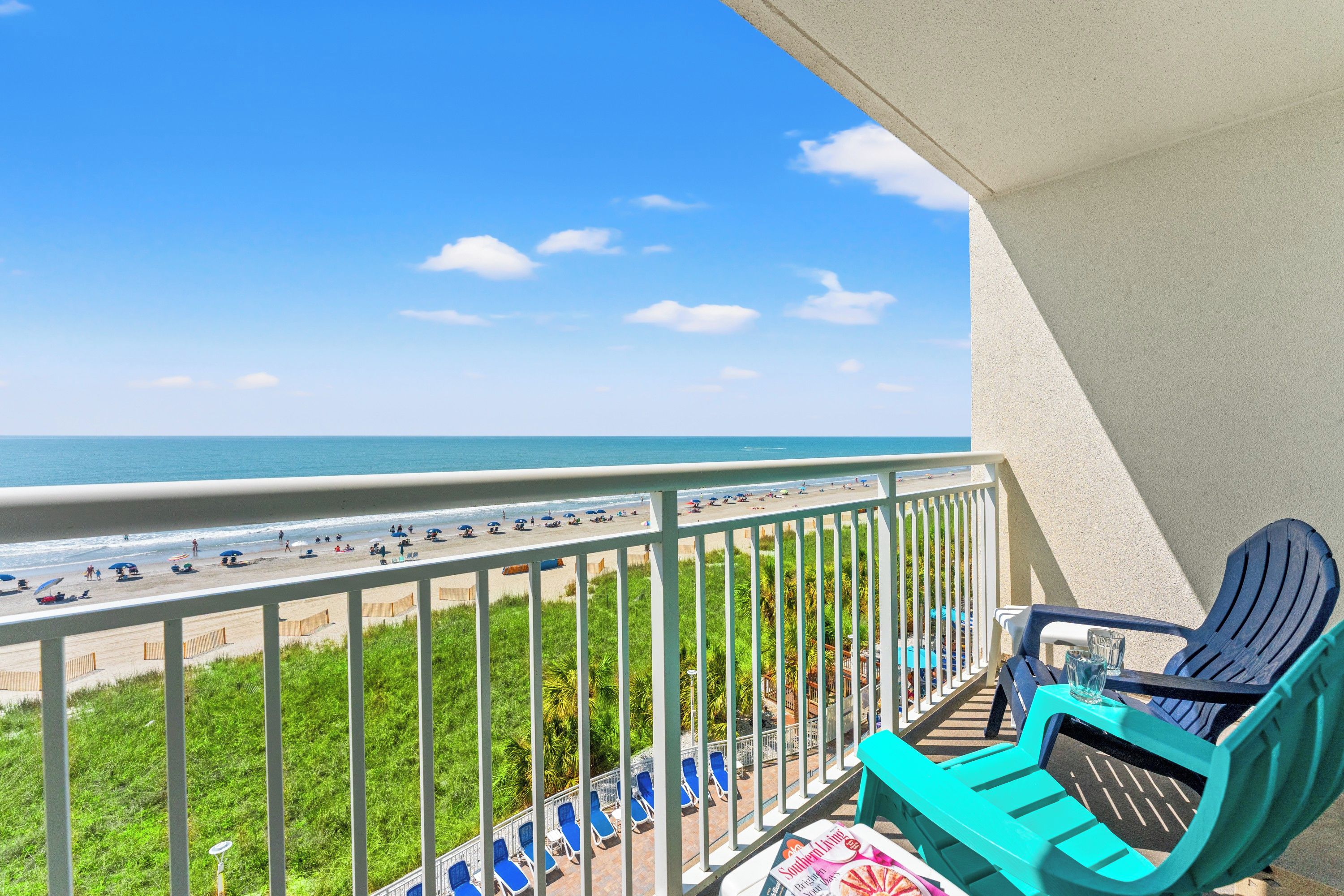 A balcony with a view of a beach, a pool area with lounge chairs, and a magazine on the table.