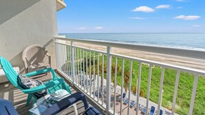 Een balkon met turquoise stoelen en een tafel, met uitzicht op een strand met ligbedden en een gazon.